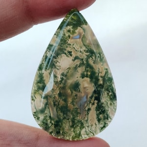 May include: A teardrop-shaped moss agate cabochon with a green, brown, and white pattern. The stone is translucent and has a smooth, polished surface.