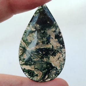 May include: A teardrop-shaped green, white, and brown moss agate cabochon. The stone has a unique and intricate pattern of moss-like inclusions.