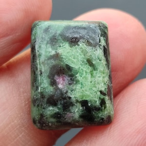 Könnte beinhalten: Ein rechteckiger grüner und schwarzer Edelstein. Der Edelstein hat eine marmorierte Optik mit dunkelgrünen, schwarzen und rosafarbenen Einschlüssen.