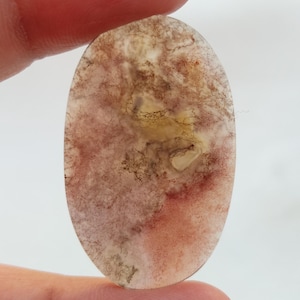 Pode incluir: Uma gema oval translúcida em cabochão com um padrão de redemoinho de marrom, rosa e branco. A pedra tem um visual natural e orgânico.