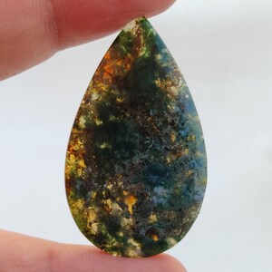 May include: A teardrop-shaped cabochon of moss agate with green, brown, and yellow patterns.