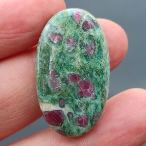 Op de afbeelding: Ovale cabochon edelsteen met een groene basis en roze insluitsels. De steen heeft een gemarmerde uitstraling met een mix van licht- en donkergroene tinten. De roze insluitsels zijn verspreid over de hele steen en lijken kleine, onregelmatige vormen te hebben.