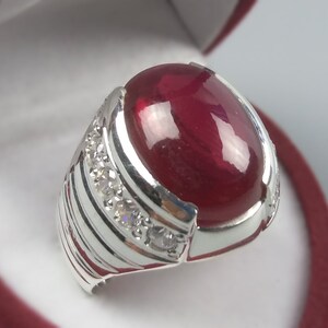 May include: A silver ring featuring a large, oval-shaped, deep red gemstone. The ring's band has a ribbed design on the sides and is accented with small, clear gemstones. The setting is polished and reflective.