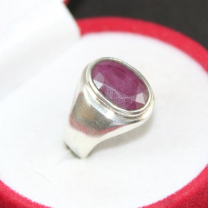 May include: A silver ring featuring a large, oval-shaped, faceted ruby gemstone. The ring is set in a simple, classic style.