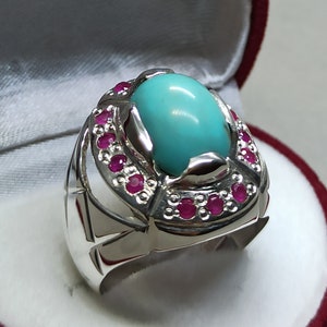 May include: A silver ring with a large turquoise stone set in a silver bezel. The ring is surrounded by a row of small red stones.