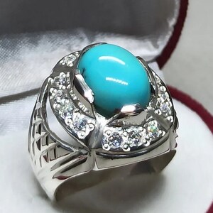 May include: A silver ring with a large turquoise stone set in the center. The ring is surrounded by smaller white stones.