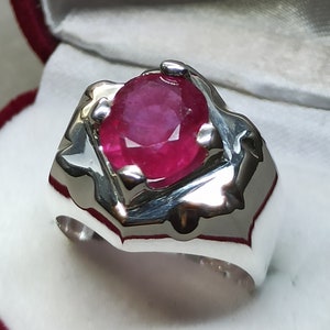 Może przedstawiać: Srebrny pierścionek z dużym, owalnym rubinem. Rubin jest intensywnie czerwony i osadzony w srebrnej obrączce z ozdobnym wzorem.