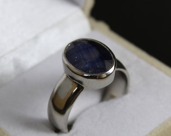 May include: A silver ring with a large, oval, dark blue gemstone. The gemstone is set in a bezel setting, and the band is smooth and polished. The ring is displayed in a white jewelry box.
