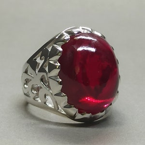 May include: A silver ring with a large, oval, deep red gemstone. The ring's band features an openwork design. The gemstone is set in a bezel setting, and the ring is displayed against a light gray background.