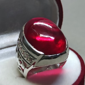 May include: A silver ring featuring a large, oval-shaped, red gemstone set in the centre. The ring is surrounded by smaller, clear gemstones.