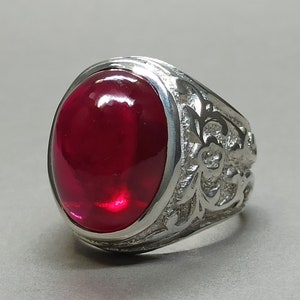 May include: A silver ring with a large, oval-shaped, red gemstone set in the center. The band of the ring is decorated with an intricate floral design.