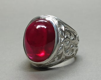 May include: A silver ring with a large, oval-shaped, red gemstone set in the center. The band of the ring is decorated with an intricate floral design.