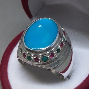 May include: A silver ring with a large, oval, bright turquoise stone as the centerpiece. The ring is accented with small red and green gemstones around the turquoise stone. The ring is set in a velvet-lined box.