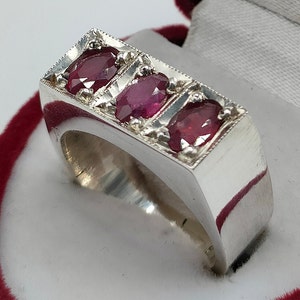 May include: A silver ring with three red ruby gemstones set in a rectangular band.
