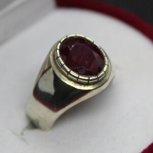 May include: A silver ring featuring a large, oval-shaped ruby gemstone set in a silver bezel. The ruby is a deep red colour and has a faceted cut.