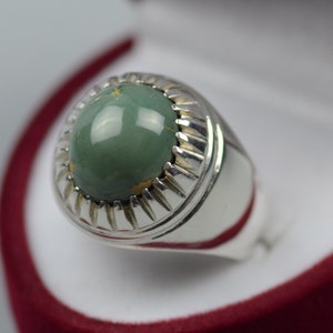 May include: A silver ring with a green gemstone set in a decorative silver bezel. The ring is displayed in a red velvet box.