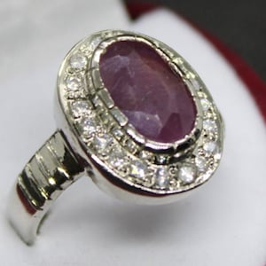 May include: A silver ring featuring a large oval pink gemstone, encircled by smaller white gemstones.