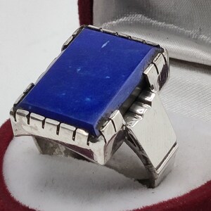 May include: A silver ring with a large, rectangular, dark blue lapis lazuli stone set in a detailed silver bezel. The ring is in a red velvet ring box.