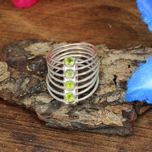 May include: A silver ring with a unique design, featuring multiple thin bands and a central rectangular section. The rectangular section holds four round, bright green gemstones. The ring is displayed on a piece of textured wood.