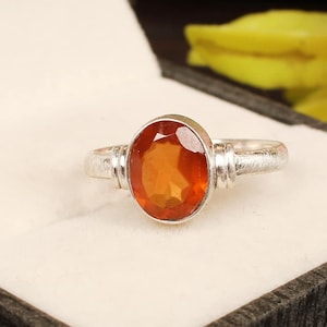 May include: A silver ring with an oval-shaped orange gemstone. The ring is set in a simple band with a textured finish.
