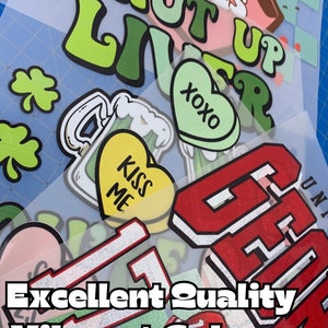 May include: A collection of iron-on heat transfer vinyl designs featuring various phrases and images, including "Love Bites Shut Up Liver", "XOXO", "Kiss Me", and a green shamrock. The designs are ready to be pressed onto fabric.