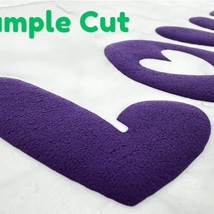 May include: A close-up of a purple felt "Love" cutout with a heart shape in the center. The text "Sample Cut" is visible in the top left corner of the image.