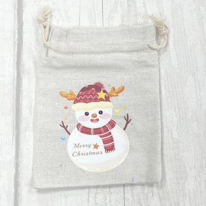May include: A small, beige burlap gift bag with a snowman design. The snowman is wearing a red and white hat, a red and white scarf, and has a yellow star on its hat. The bag says "Merry Christmas" in brown text.