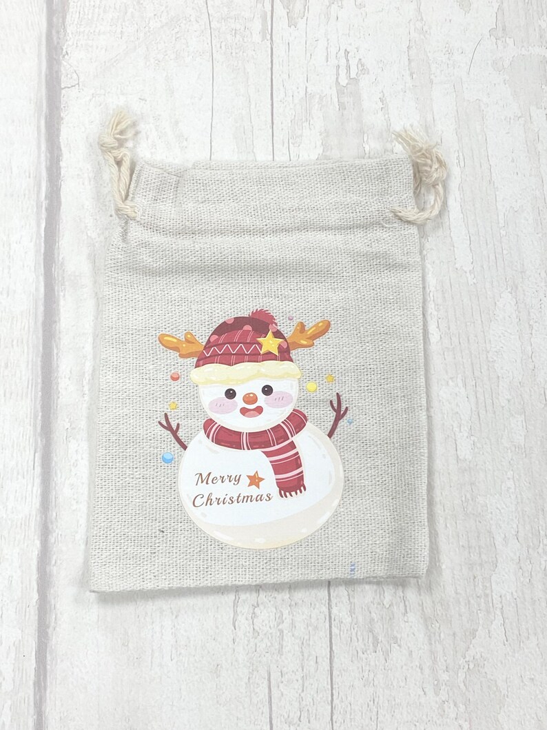 May include: A small, beige burlap gift bag with a snowman design. The snowman is wearing a red and white hat, a red and white scarf, and has a yellow star on its hat. The bag says "Merry Christmas" in brown text.