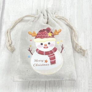 May include: A small, beige hessian gift bag with a drawstring closure. The bag features a printed design of a snowman wearing a red and white hat and scarf. The snowman has a star on its hat and the text "Merry Christmas" is printed below the snowman.
