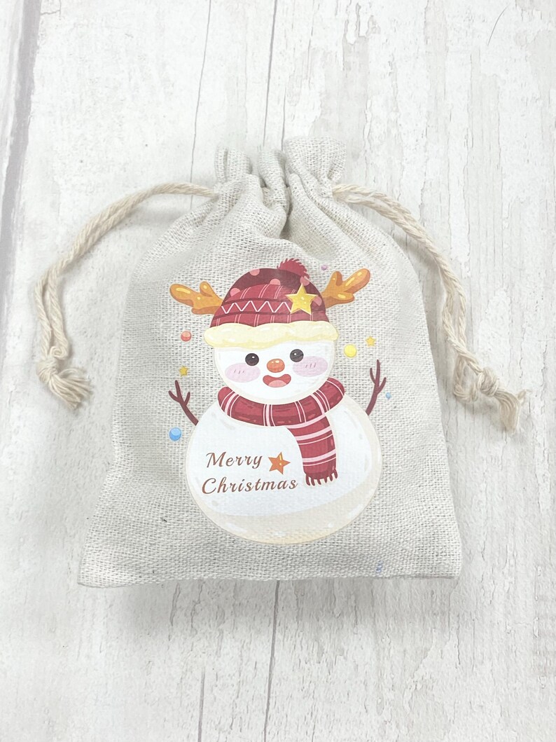 May include: A small, beige hessian gift bag with a drawstring closure. The bag features a printed design of a snowman wearing a red and white hat and scarf. The snowman has a star on its hat and the text "Merry Christmas" is printed below the snowman.