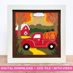 May include: A framed paper art piece featuring a red pickup truck carrying two orange pumpkins. The scene includes a red barn, trees with orange and yellow leaves, and a yellow road. The frame is white.