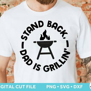 May include: White t-shirt with black text that reads "Stand Back Dad Is Grillin'" with a grill and flames in the center.