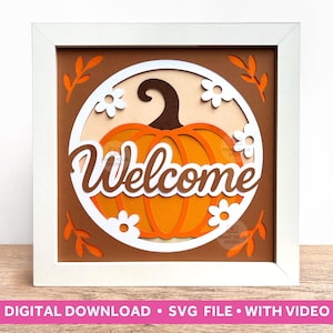 May include: A layered paper art piece in a white frame. The design features an orange pumpkin with the word "Welcome" in cursive. White flowers and orange leaves surround the pumpkin on a brown background. The frame is on a wooden surface.