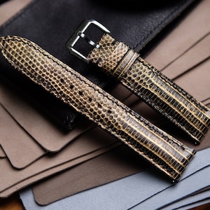 May include: Two brown leather watch straps with a textured pattern. The straps have a silver buckle and are laid out on a stack of brown and gray fabric.