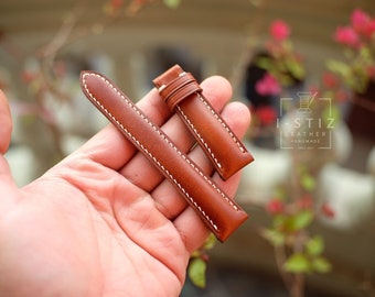 May include: A pair of brown leather watch straps with white stitching. The straps are held in a hand, showcasing the detailed stitching and the quality of the leather. The watch straps are a warm brown color.