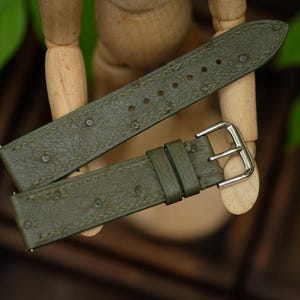 Pode incluir: Pulseira de relógio verde-oliva com superfície texturizada e fivela prateada. A pulseira é feita de um material que se assemelha ao couro de avestruz. A pulseira do relógio é mostrada em um fundo desfocado com folhas verdes.