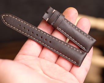 May include: Two brown leather watch straps with white stitching. The straps are laid out on a hand, showing the stitching detail.