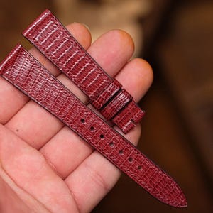 以下が含まれることがあります： トカゲ革のような型押しが施された、深みのある赤色の時計ベルト。ベルトは2つのパーツに分かれており、長い方のパーツには調整用の穴が並び、短い方のパーツには2つのループが付いています。