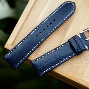 May include: Two dark blue leather watch straps with white stitching. The straps have a silver buckle and are laid out on a wooden surface.