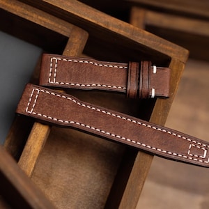 May include: Two brown leather watch straps with white stitching are displayed in a wooden box. The straps have a textured appearance and are designed for a watch. A business card with the text "STIZ LEATHER" is also visible.