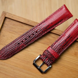 May include: Two red lizard skin watch straps with silver buckles. The straps are laid out on a wooden surface.