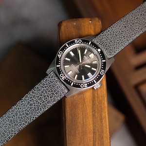 May include: A silver watch with a black face and a gray leather band. The watch has a black bezel with white markings and a date window at the 3 o'clock position.
