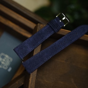 Può includere: Un cinturino per orologio in camoscio blu scuro con finitura testurizzata e fibbia in metallo color argento. Il cinturino è composto da due pezzi, uno con fibbia e passante, e l'altro con un'estremità affusolata, per il fissaggio all'orologio.