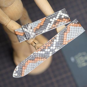 May include: Two watch straps with a snakeskin pattern in gray, white, and orange are displayed. The straps are made of a textured material and are positioned on a wooden mannequin. The background is a dark surface.