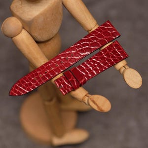 Puede incluir: Una correa de reloj roja brillante con un patrón texturizado de piel de cocodrilo. La correa consta de dos piezas separadas, una con un extremo puntiagudo y varios orificios de ajuste, y la otra con una zona de hebilla y una trabilla deslizante.