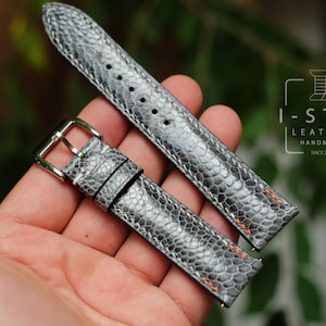 Puede incluir: Dos correas de reloj de cuero gris con costuras naranjas. Las correas tienen un patrón texturizado que se asemeja a la piel de serpiente. Las correas están dispuestas sobre una mano.