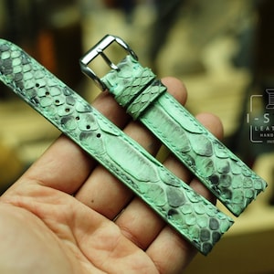May include: A pair of watch straps made of green and black snakeskin leather. The straps have a silver buckle and are handmade. The I-STIZ LEATHER logo is visible in the background.