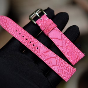 May include: A pink leather watch strap with a textured, alligator-like pattern. The strap has white stitching and a silver buckle.