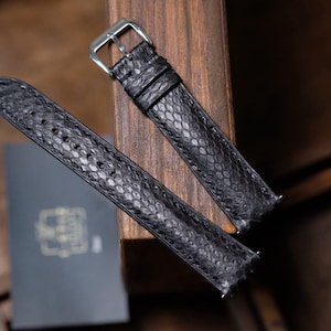 May include: A black watch strap featuring a textured snakeskin pattern with a silver-toned metal buckle. The strap consists of two pieces, one with a series of punched holes and the other with a metal pin buckle and two leather keepers.