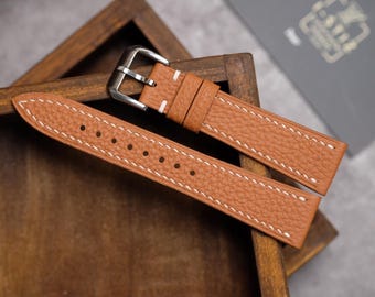 May include: A tan leather watch strap with white stitching and a silver buckle. The strap is displayed on a wooden surface, showcasing its texture and design. The watch band is new.